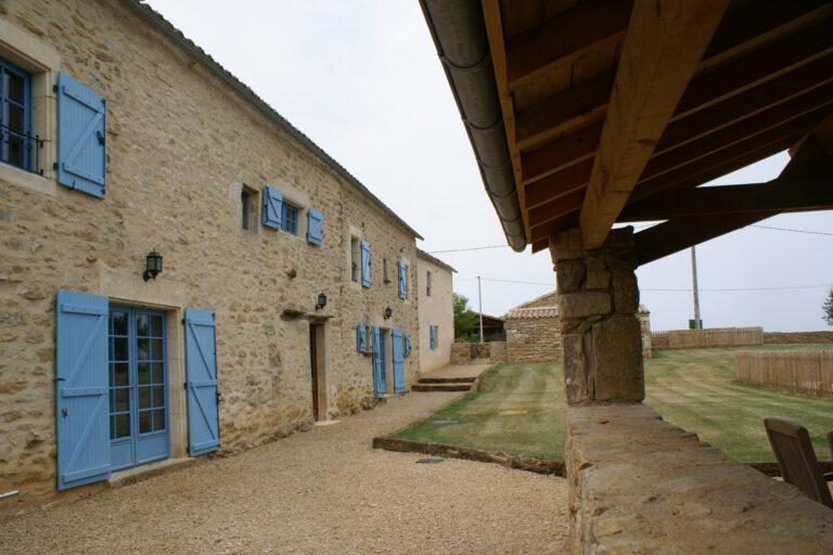 The farmhouse frontage in stone with pale blue shutters, seen from beneath the barn roof